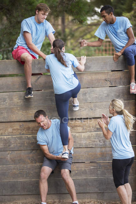 Партнеры по команде помогают женщине перелезть через стену на полосе препятствий в учебном лагере — стоковое фото