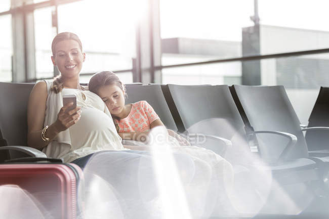 Schwangere Mutter mit Handy und schlafender Tochter im Abflugbereich des Flughafens — Stockfoto
