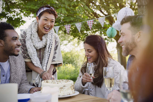 Lächelnde Freunde, die Champagner trinken und Geburtstagstorte am Terrassentisch anschneiden — Stockfoto