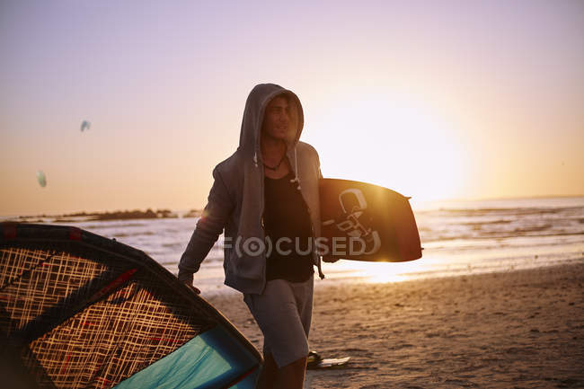 Mann im Kapuzenpulli trägt Kiteboard-Ausrüstung am Strand von Sonnenuntergang — Stockfoto