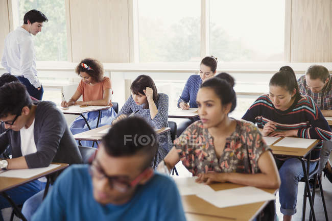 Профессор наблюдает за студентами, сдающими экзамены за партами в классе — стоковое фото