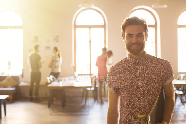 Portrait smiling casual businessman with skateboard in sunny open office — Stock Photo