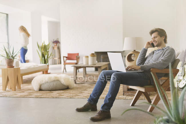Homem falando no celular e usando laptop na sala de estar — Fotografia de Stock