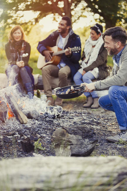 Amigos tocando guitarra e cozinhar cachorros-quentes na fogueira — Fotografia de Stock