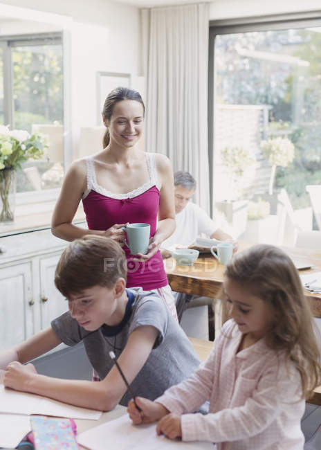 Мать с кофе наблюдает, как дочь и сын делают домашнюю работу — стоковое фото