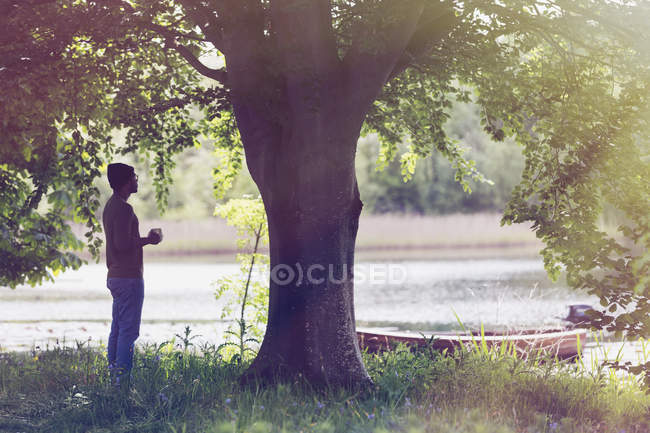 Uomo che beve caffè sotto l'albero al lago idilliaco — Foto stock