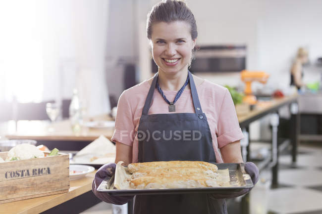 Portrait smiling woman holding tray of food in cooking class kitchen — Stock Photo