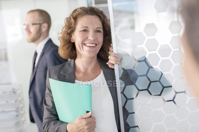 Портрет женщины с документами, стоящей у стеклянной двери, коллега на заднем плане — стоковое фото