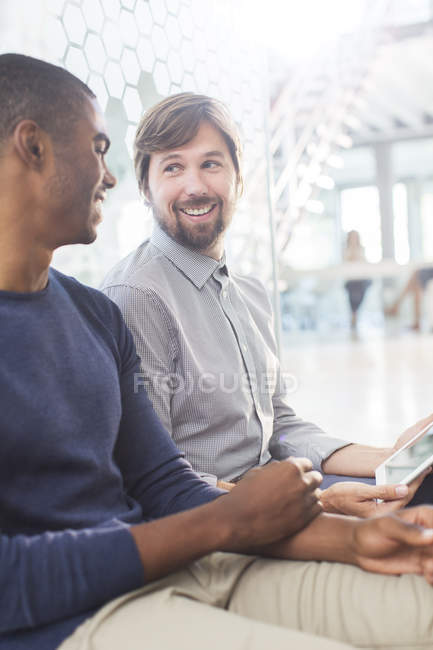 Двое улыбающихся мужчин разговаривают, держат цифровые планшеты в офисном коридоре — стоковое фото