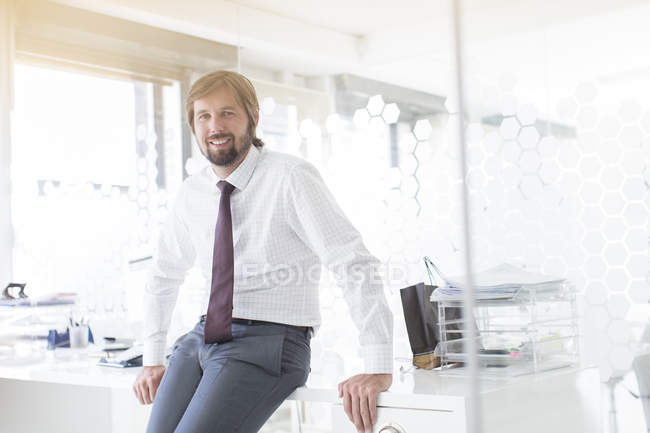 Porträt eines lächelnden Geschäftsmannes mit Hemd und Krawatte, der im Büro am Schreibtisch lehnt — Stockfoto