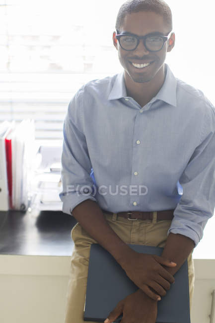 Portrait of smiling young man wearing glasses and blue shirt leaning on desk in office — Stock Photo