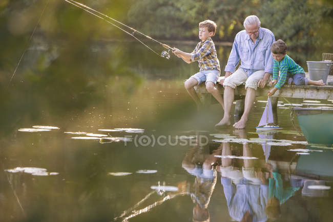 Дедушка и внуки рыбачат и играют с игрушечным парусником на озере — стоковое фото