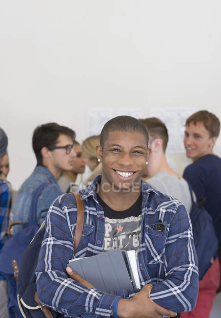 Männlicher Student hält Bücher in der Hand und lächelt mit anderen Studenten im Hintergrund in die Kamera — Stockfoto