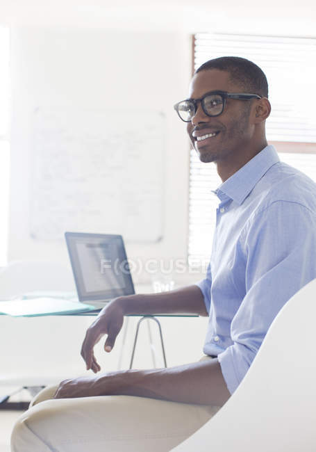Retrato de jovem sorridente vestindo óculos e camisa azul sentado na mesa com laptop — Fotografia de Stock