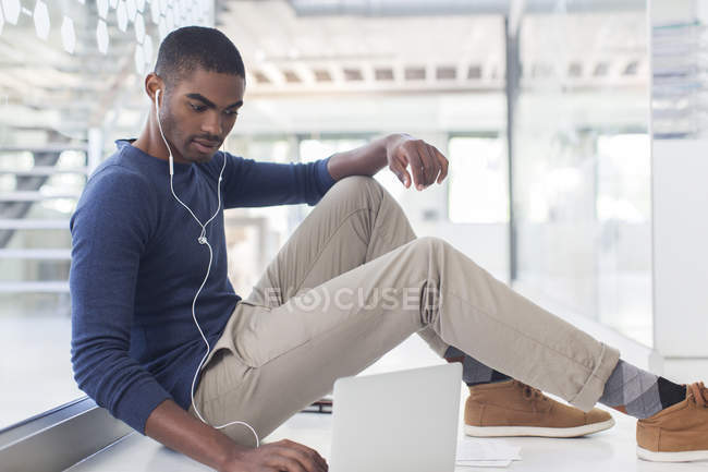 Retrato de homem de negócios sentado no chão no escritório moderno, usando laptop e fones de ouvido — Fotografia de Stock