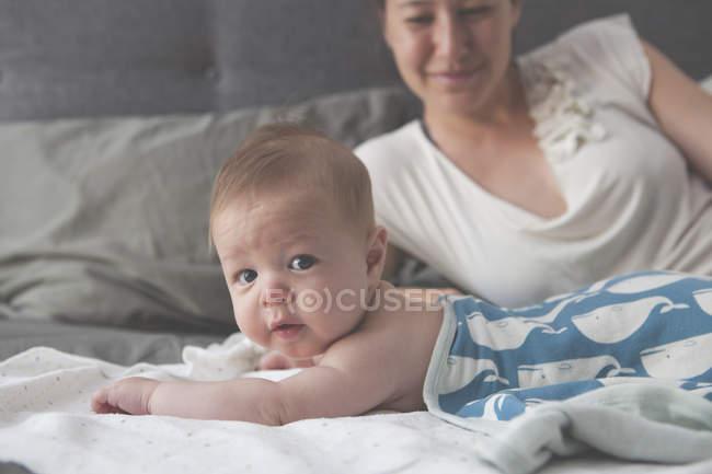 Портрет малыша, лежащего на кровати с улыбкой матери на заднем плане — стоковое фото