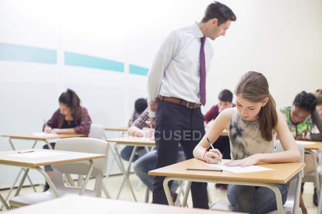 Profesor masculino que supervisa a estudiantes que escriben su examen GCSE en el aula - foto de stock