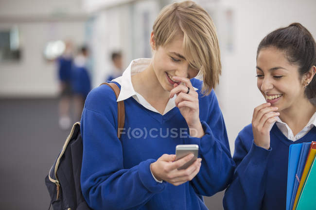 Dos estudiantes sonrientes mirando el teléfono móvil en el pasillo de la escuela - foto de stock