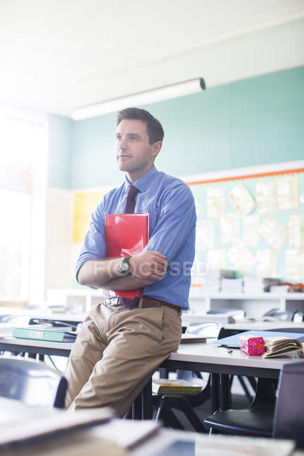 Retrato del profesor varón apoyado en el escritorio en el aula - foto de stock