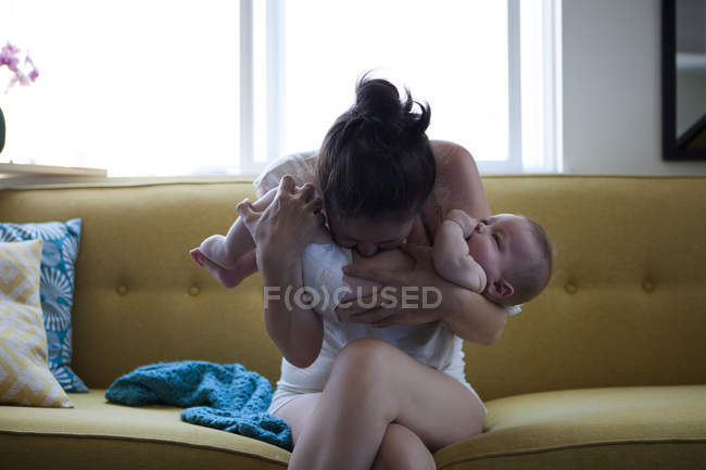 Madre che tiene e bacia la pancia del bambino, seduta sul divano alla finestra — Foto stock