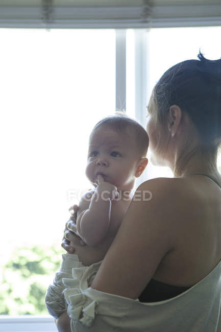 Madre che tiene in braccio il bambino e guarda attraverso la finestra — Foto stock