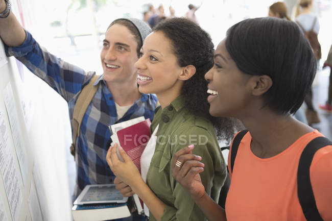 Three smiling students looking at information board, holding books and digital tablet — Stock Photo