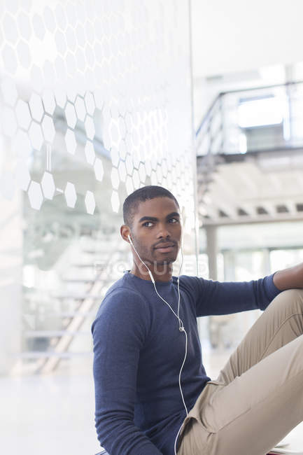 Hombre escuchando música en los auriculares, sentado en el suelo en el pasillo de la oficina - foto de stock