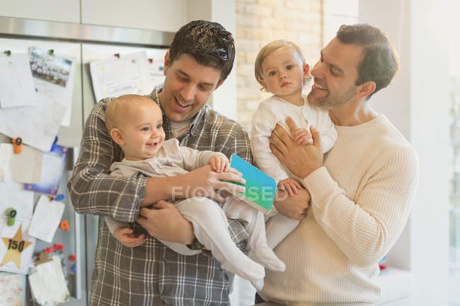 Male gay parents holding baby sons in kitchen — Stock Photo