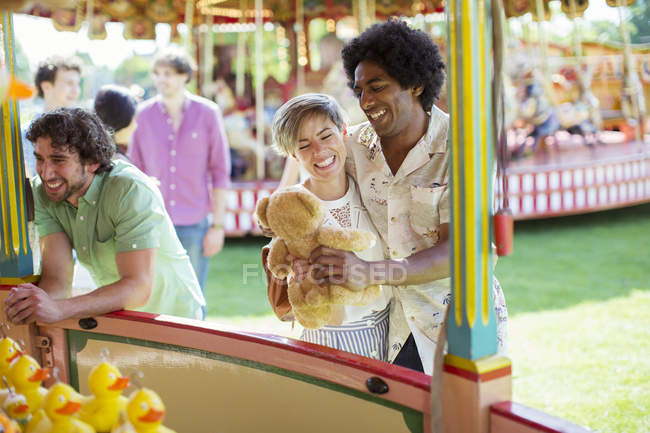 Smiling couple holding teddy bear next to fishing game in amusement park — Stock Photo