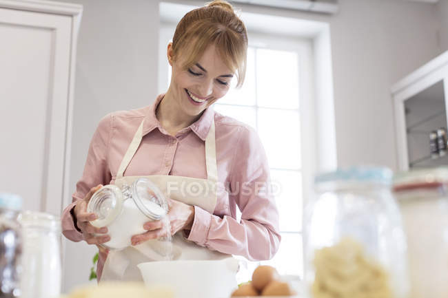 Mulher sorrindo assar, derramando açúcar em tigela na cozinha — Fotografia de Stock
