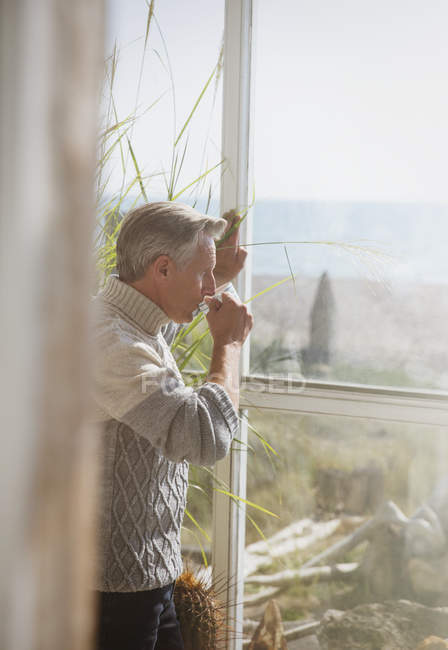 Старший мужчина пьет кофе у окна солнечного пляжного домика — стоковое фото