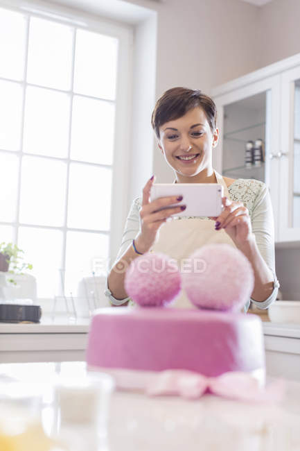 Женщина-повар с камерой телефона фотографирует розовый свадебный торт на кухне — стоковое фото