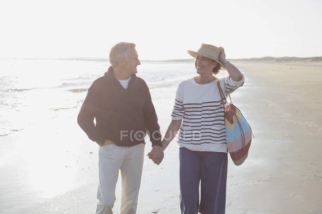 Sonriendo pareja madura cogida de la mano y caminando en la playa soleada - foto de stock