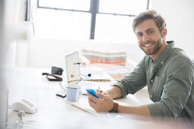 Portrait of young man sitting at desk with mobile phone and laptop — Stock Photo