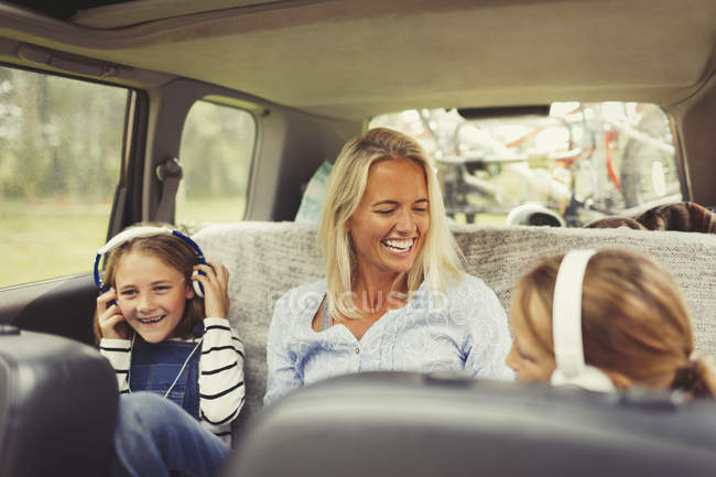Riendo madre e hijas con auriculares en el asiento trasero del coche - foto de stock