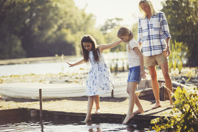 Mother watching daughters sticking toes in water on lakeside dock — Stock Photo