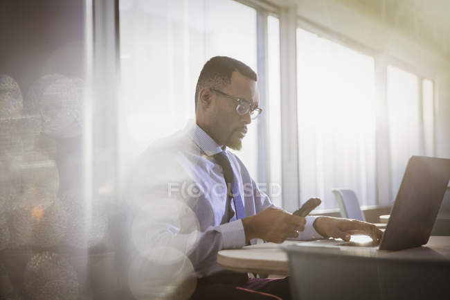 Grave uomo d'affari che scrive con il cellulare al computer portatile in ufficio — Foto stock