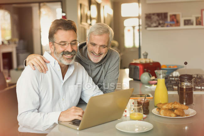 Couple gay masculin utilisant un ordinateur portable et manger le petit déjeuner au comptoir de la cuisine — Photo de stock