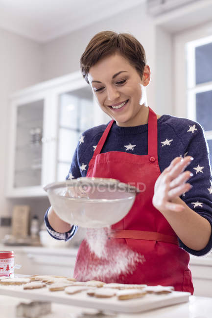 Mujer sonriente horneando, tamizando azúcar sobre galletas en la cocina - foto de stock