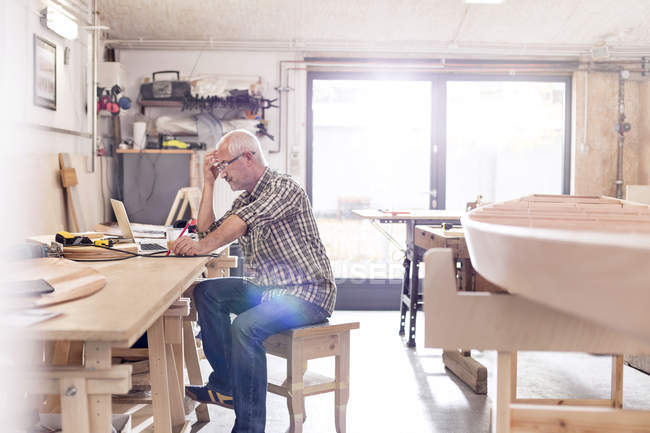 Tischler arbeitet am Laptop auf Werkbank neben Holzboot in Werkstatt — Stockfoto