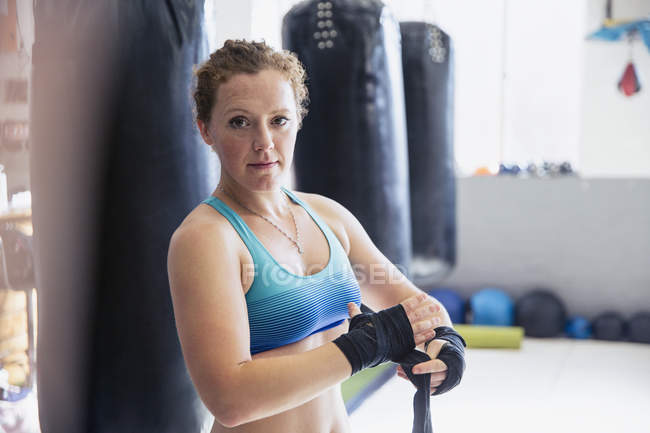 Portrait confident, tough female boxer wrapping wrists next to punching bags in gym — Stock Photo