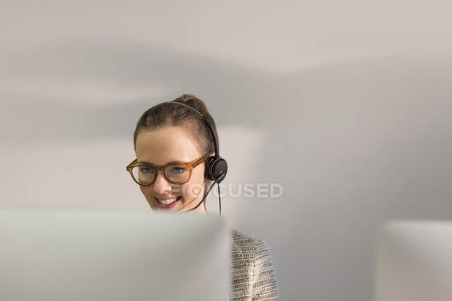 Smiling female telemarketer wearing headset talking on telephone at computer in office — Stock Photo