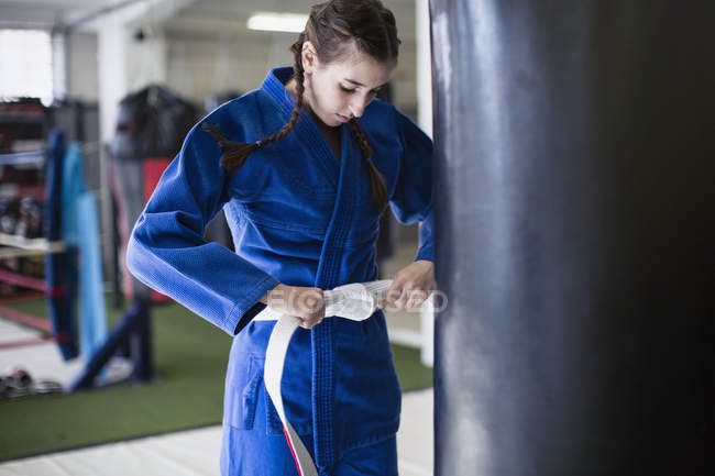 Молодая женщина затягивает пояс дзюдо у боксерской груши в спортзале — стоковое фото