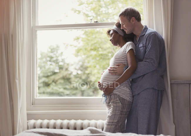 Affectionate pregnant couple in pajamas hugging at window — Stock Photo