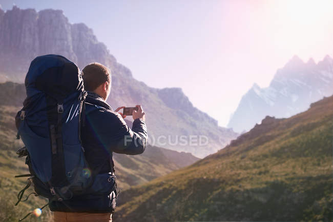 Jovem com mochila usando telefone câmera no vale ensolarado abaixo das montanhas — Fotografia de Stock