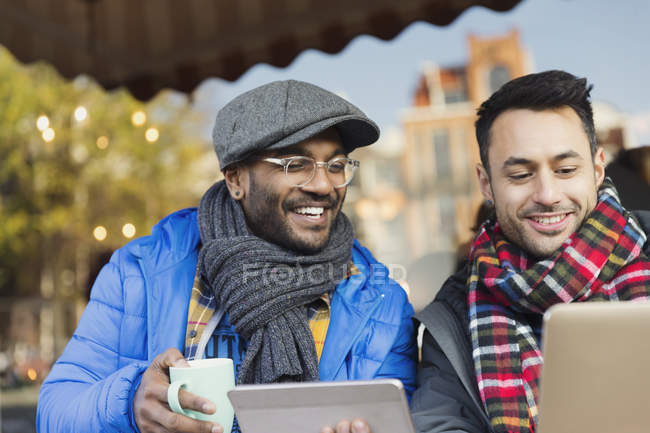 Smiling young men friends drinking coffee and using laptop and digital tablet at sidewalk cafe — Stock Photo