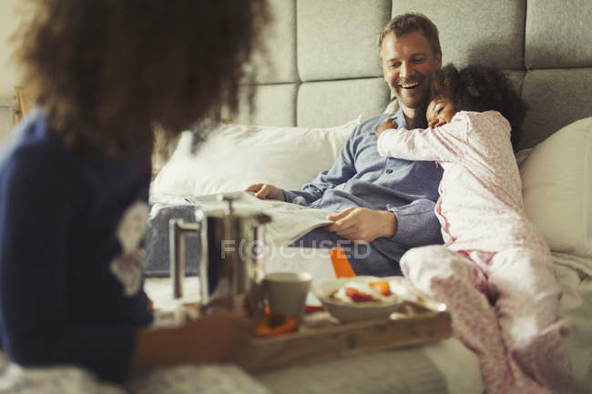 Hija multiétnica abrazando padre leyendo periódico en la cama - foto de stock