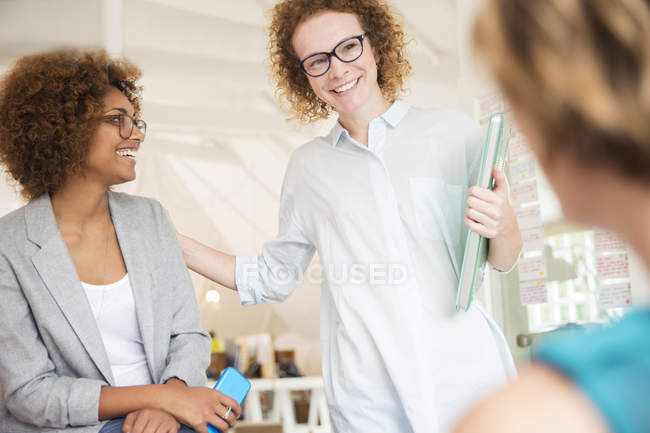 Mujeres sonriendo y hablando en la oficina, sosteniendo el teléfono inteligente y el ordenador portátil - foto de stock