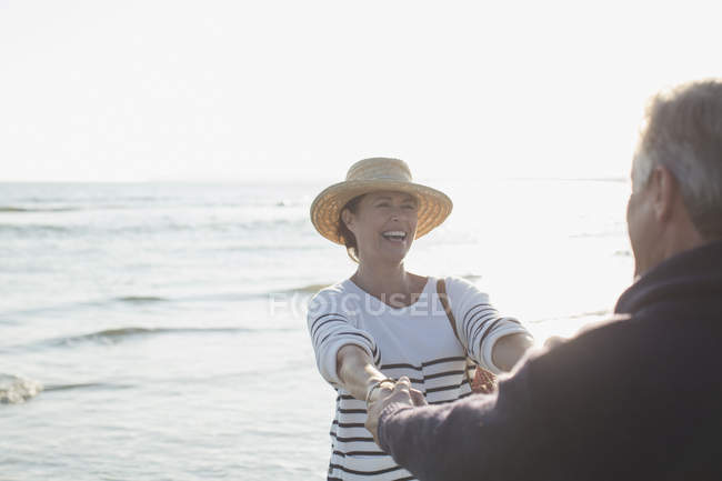 Playful mature couple holding hands on sunny ocean beach — Stock Photo