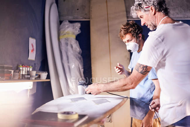 Männliche Surfbrettdesigner mit Schutzmasken schleifen Surfbrett in Werkstatt — Stockfoto